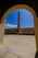 A tall, narrow chimney or smokestack is centrally framed by an arched doorway. The scene includes a courtyard with a stone or concrete floor and walls surrounding the chimney. The sky is clear and bright blue, adding contrast to the structure's earthy tones.