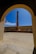 A tall, narrow chimney or smokestack is centrally framed by an arched doorway. The scene includes a courtyard with a stone or concrete floor and walls surrounding the chimney. The sky is clear and bright blue, adding contrast to the structure's earthy tones.