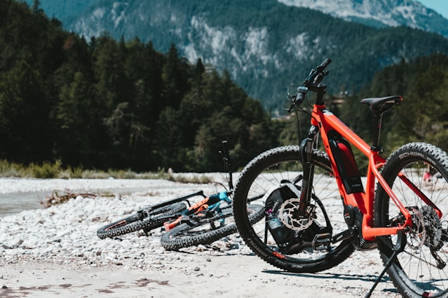 Two mountain bikes rest on a rocky, sandy terrain with forested mountains in the background. The prominent bicycle is brightly painted in red and black, equipped with thick tires, and appears to be an electric model. The landscape suggests an adventurous outdoor setting with vibrant greenery and rugged rocks.