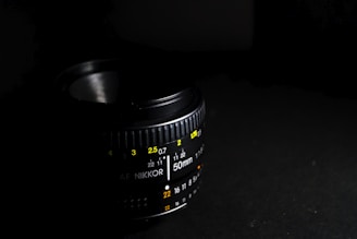 A close-up of a camera lens showcasing various aperture and focus markings in yellow and white colors against a black background. The lens is partially lit, creating a dramatic contrast between light and shadow.