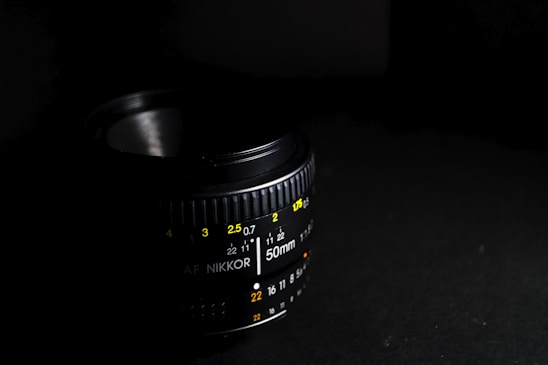 A close-up of a camera lens showcasing various aperture and focus markings in yellow and white colors against a black background. The lens is partially lit, creating a dramatic contrast between light and shadow.