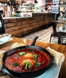 In a cozy cafe setting, a cast iron skillet filled with shakshuka is placed on a wooden table. The dish features two poached eggs in a rich tomato sauce, garnished with slices of jalape&ntilde;o and cilantro. Surrounding the pan are slices of crusty bread, with a side dish containing a small salad. In the background, a rustic counter with a variety of kitchen utensils and food items adds to the inviting atmosphere.