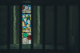 A vertically aligned stained glass window is framed by dark bars in the foreground, creating a sense of separation. The window features vibrant patterns and shapes in various colors, including red, blue, green, and yellow, set against a darker background. The surrounding walls appear aged and textured, adding a rustic charm.