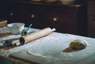 A rustic kitchen setting with a dough ball on a floured countertop. A wooden rolling pin rests nearby alongside baking utensils such as a metal cutter and a measuring cup. The background includes a floral-patterned tablecloth and wooden furniture, evoking a traditional and cozy atmosphere.