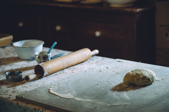 A rustic kitchen setting with a dough ball on a floured countertop. A wooden rolling pin rests nearby alongside baking utensils such as a metal cutter and a measuring cup. The background includes a floral-patterned tablecloth and wooden furniture, evoking a traditional and cozy atmosphere.