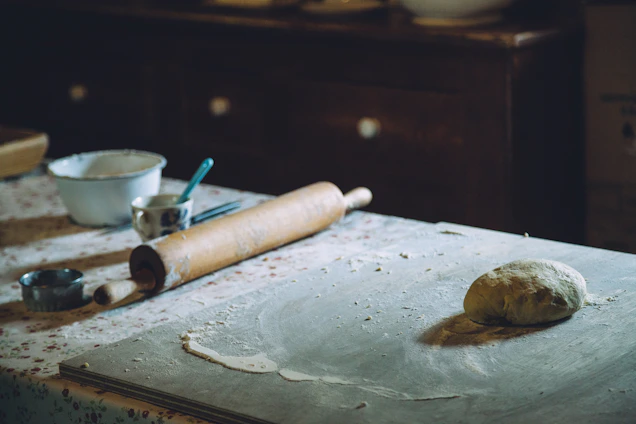 A rustic kitchen scene showing a pair of hands rolling fresh dough with traditional tools, surrounded by natural ingredients.