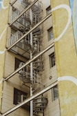 A spiral metal staircase is attached to the exterior of a beige building. The building features several windows and is partially framed by a wall with yellow and white abstract patterns.