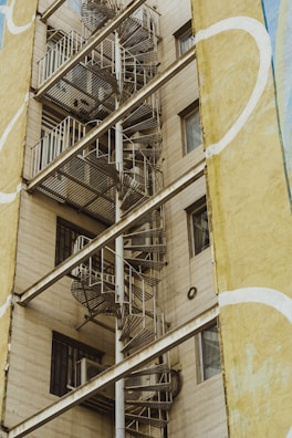 A spiral metal staircase is attached to the exterior of a beige building. The building features several windows and is partially framed by a wall with yellow and white abstract patterns.