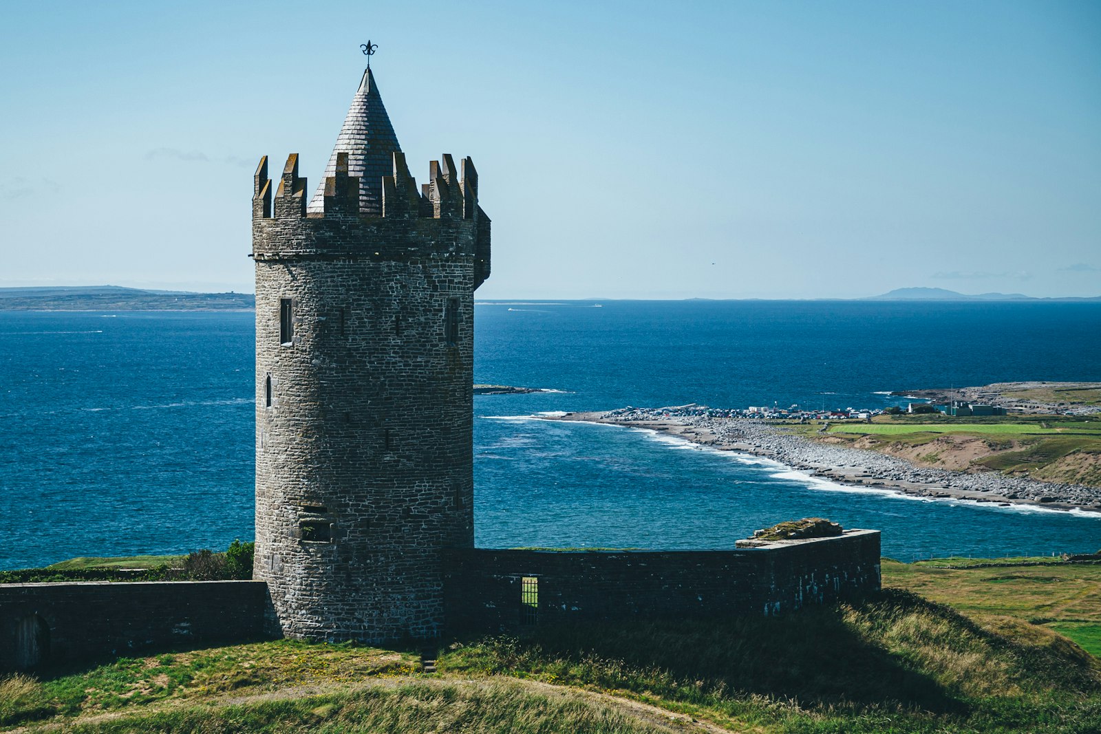 Round grey stone tower house with a conical roof standing on a green hill above a rocky shoreline and calm blue sea