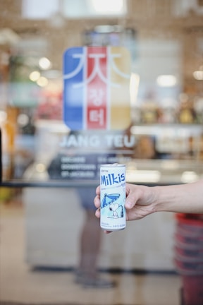 A hand is holding a can of Milkis, a Korean carbonated milk beverage, in front of a glass door with a colorful sign and the phrase 'JANG TEU' on it.
