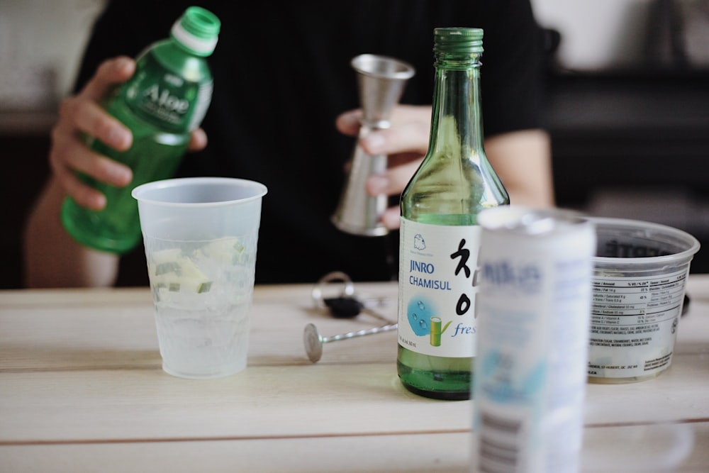 person holding bottle beside table with cup, bottle, and can