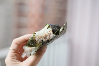 A hand holding a partially eaten rice ball wrapped in seaweed, revealing a filling, possibly tuna. The background is blurred, focusing attention on the snack.