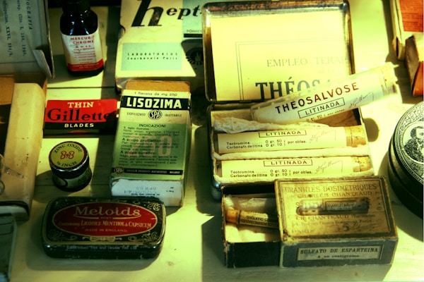 An assortment of vintage pharmaceutical and grooming products are arranged on a surface. Items include a box of Gillette razor blades, a small bottle labeled Mercurochrome, various tubes marked with names like Lizinada and Theosalvose, and tins for items such as Meloids and zinc oxide ointment. Packaging features old-fashioned fonts and design, with diverse languages visible on labels.