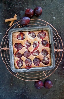 A square baking tray holds a freshly baked plum cake with visible pieces of plum embedded in the golden-brown crust. The tray is placed on a circular wire rack on a dark countertop. Surrounding the cake are whole plums and a couple of cinnamon sticks, enhancing the rustic and homemade feel.