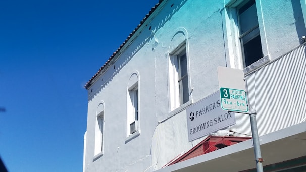 A two-story building with a light blue exterior featuring arched windows and a textured facade. The building houses 'Parker's Grooming Salon,' as indicated by a sign with paw prints. In front of the building, a parking sign specifies a 3-hour parking limit from 9 a.m. to 6 p.m. The sky is clear and vibrantly blue.