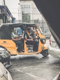 A yellow auto-rickshaw is parked on a street, occupied by several passengers. The street appears wet, suggesting recent rainfall. There are various cars nearby, and buildings visible in the background. The scene has an urban setting with a residential or commercial area in view.