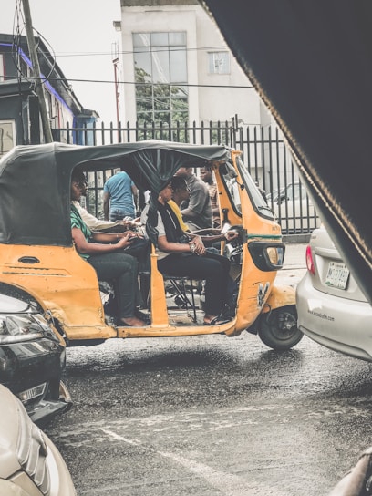 A yellow auto-rickshaw is parked on a street, occupied by several passengers. The street appears wet, suggesting recent rainfall. There are various cars nearby, and buildings visible in the background. The scene has an urban setting with a residential or commercial area in view.