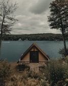 An A-frame cabin sits at the edge of a serene lake, surrounded by lush greenery and tall trees under an overcast sky. The water is calm, and a distant boat creates a small wake near the far shoreline where additional homes are visible amidst the dense forest.