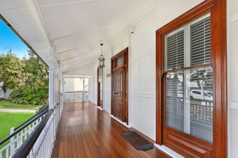 A polished wooden porch lined with white railings and walls, featuring a dark wood front door. There's a metal lantern hanging from the ceiling, and a view of a green lawn and trees is visible in the background.