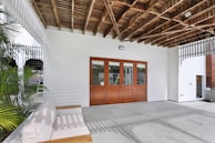 A covered outdoor patio area with a wooden ceiling and white paneling on the walls. Natural light filters through, casting shadows on the stone tile floor. There's a cushioned bench with wooden accents on one side and large wooden-framed glass doors leading inside. Green plants decorate the area, adding a touch of nature.