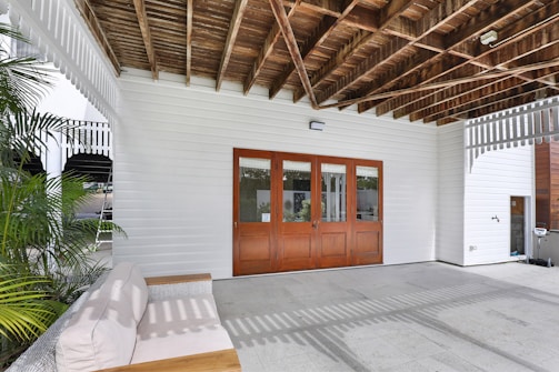 A covered outdoor patio area with a wooden ceiling and white paneling on the walls. Natural light filters through, casting shadows on the stone tile floor. There's a cushioned bench with wooden accents on one side and large wooden-framed glass doors leading inside. Green plants decorate the area, adding a touch of nature.