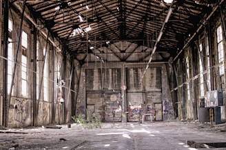 An abandoned industrial building with a decaying interior. The roof is partially collapsed, allowing light to filter through broken panels. Graffiti covers the walls, and debris is scattered across the floor. Large windows line the sides, though many are broken. In the center, there is a barrier with more graffiti, and plants have begun to grow amidst the ruin.