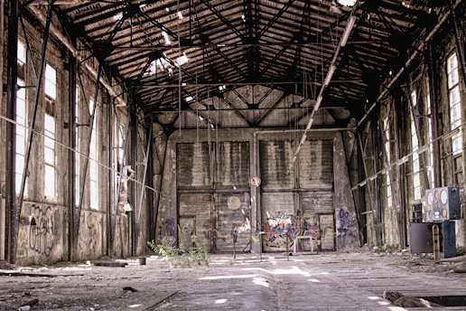 An abandoned industrial building with a decaying interior. The roof is partially collapsed, allowing light to filter through broken panels. Graffiti covers the walls, and debris is scattered across the floor. Large windows line the sides, though many are broken. In the center, there is a barrier with more graffiti, and plants have begun to grow amidst the ruin.