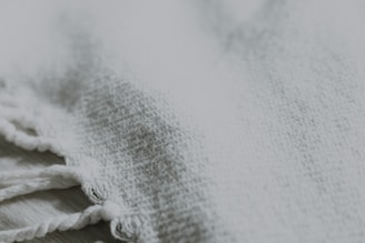 A close-up of a textured fabric with a woven pattern. The image prominently features a fringe with twisted tassels on one side, showcasing the intricate detail of the material.