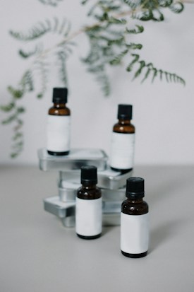 Four small amber glass bottles with black caps are arranged among several rectangular, metal tins on a solid surface. Delicate green fern fronds hang above, partially out of focus, creating an organic backdrop.