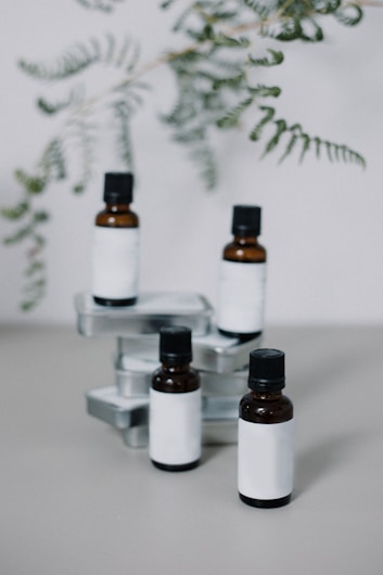 Four small amber glass bottles with black caps are arranged among several rectangular, metal tins on a solid surface. Delicate green fern fronds hang above, partially out of focus, creating an organic backdrop.