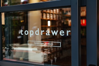 The image features a glass storefront panel with the text 'topdrawer' and 'TOOLS FOR NOMADS' written on it. Behind the glass, shelves filled with various items are visible, suggesting a store interior. The overall aesthetic is modern and clean.