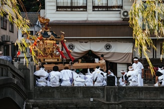 A group of people dressed in white traditional clothing gathers around a decorative portable shrine, known as a mikoshi, in a cultural setting. The shrine is ornately adorned with gold and red embellishments. The scene is set outdoors in front of a building with traditional architecture, and branches hang down into the frame.