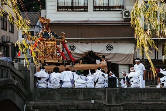 A group of people dressed in white traditional clothing gathers around a decorative portable shrine, known as a mikoshi, in a cultural setting. The shrine is ornately adorned with gold and red embellishments. The scene is set outdoors in front of a building with traditional architecture, and branches hang down into the frame.