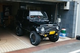 A black off-road vehicle, possibly a Suzuki, is parked in a tiled garage area. It has large tires with rugged tread and a visible winch at the front. There are some items like a green bag and a box to the right. The garage has a dark tiled wall and a few bicycles are visible in the background.