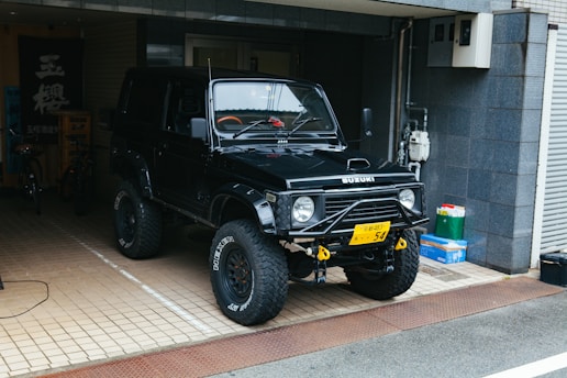 A black off-road vehicle, possibly a Suzuki, is parked in a tiled garage area. It has large tires with rugged tread and a visible winch at the front. There are some items like a green bag and a box to the right. The garage has a dark tiled wall and a few bicycles are visible in the background.