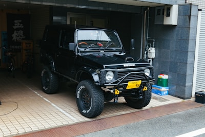 A black off-road vehicle, possibly a Suzuki, is parked in a tiled garage area. It has large tires with rugged tread and a visible winch at the front. There are some items like a green bag and a box to the right. The garage has a dark tiled wall and a few bicycles are visible in the background.
