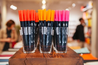 A collection of black markers with vibrant colored caps, including red, orange, and pink, are neatly organized in three transparent glass cups placed on a wooden surface. The background suggests a busy store setting with out-of-focus figures and shelves.