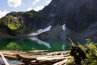 A serene mountain lake with clear, reflective water surrounded by cliffs and lush greenery. Snow patches are visible on the rocky slopes. Fallen logs rest on the shore, and the sky above is partly cloudy.