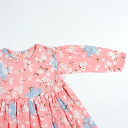 A pink floral dress featuring a pattern of white and light blue flowers with green stems. The dress has long sleeves and a gathered waist, giving it a dainty and feminine appearance.