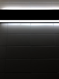 A pattern of dark tiles with a light source at the top. The tiles are arranged in a grid and have a glossy finish, reflecting some light. The light creates strong contrasts between the illuminated and darker sections.