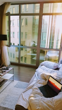 A cozy living space with a large window allowing natural light to flood in, with a view of a modern building outside. Inside, a comfortable couch draped with a white comforter hosts an open laptop displaying a sunset or sunrise scene. A small, minimalist side table contains a few items including eyeglasses and a small bowl. A floor lamp stands nearby, and a woven rug covers part of the wooden floor.