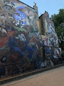 A large, colorful mural depicting a dynamic and chaotic scene that includes people in historical clothing, protestors, a police officer on horseback, and banners with political symbols. The setting appears urban with brick buildings and a streetlamp. Various elements like a chess piece, leaflets, and a diary street sign are incorporated into the composition.