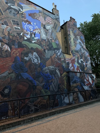 A large, colorful mural depicting a dynamic and chaotic scene that includes people in historical clothing, protestors, a police officer on horseback, and banners with political symbols. The setting appears urban with brick buildings and a streetlamp. Various elements like a chess piece, leaflets, and a diary street sign are incorporated into the composition.