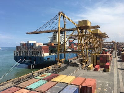 A large cargo ship docked at a busy port, surrounded by numerous colorful shipping containers stacked both on the ship and on the dock. Multiple large yellow cranes extend over the ship, used for loading and unloading the containers. The clear blue sky stretches over the calm sea, framing the industrial scene.
