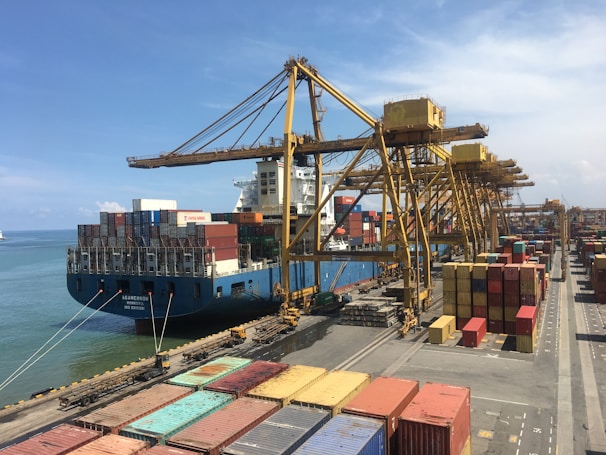 A large cargo ship docked at a busy port, surrounded by numerous colorful shipping containers stacked both on the ship and on the dock. Multiple large yellow cranes extend over the ship, used for loading and unloading the containers. The clear blue sky stretches over the calm sea, framing the industrial scene.