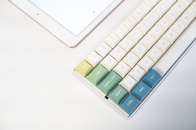 A mechanical keyboard with colorful keys is situated next to a tablet. The keys are a mix of pastel yellow, green, and blue hues, creating a calming aesthetic. The keyboard's minimalist design contrasts with the sleek surface of the tablet.