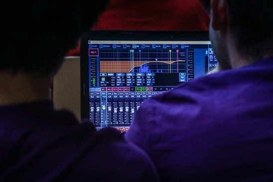 Two people are focused on a laptop displaying audio mixing software. The screen shows various controls, graphs, and faders indicating sound levels, EQ settings, and other technical details. The lighting suggests a low-light environment, contributing to an intense and concentrated atmosphere.