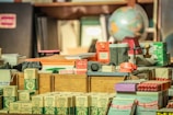 An assortment of vintage office and stationery items arranged on a desk, including boxes of crayons, a globe, stacked papers, stamps, and various labeled packages. The items have a nostalgic and old-fashioned charm, with visible text and colorful designs on the packaging.