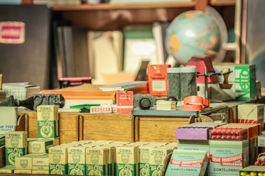 An assortment of vintage office and stationery items arranged on a desk, including boxes of crayons, a globe, stacked papers, stamps, and various labeled packages. The items have a nostalgic and old-fashioned charm, with visible text and colorful designs on the packaging.