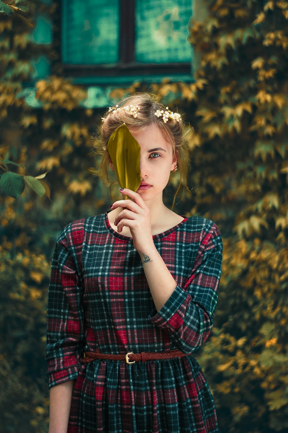 woman in green and red plaid dress standing and cover her right eye with green leaf